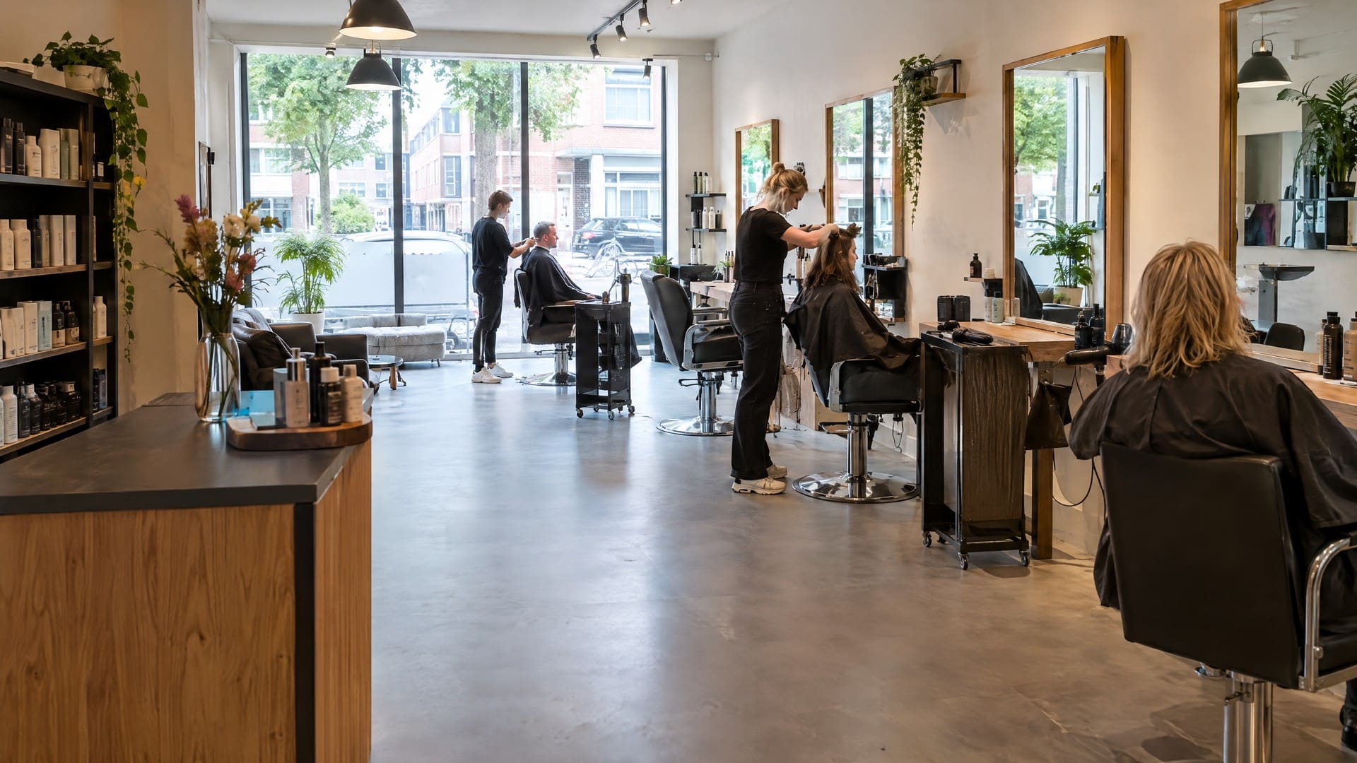 Kapsalon met stoelen voor grote spiegels, daglicht uit de etalage en planten langs de wand.